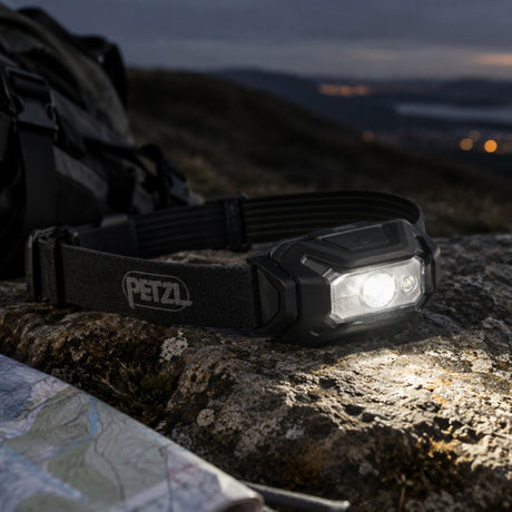 Black Petzl ARIA 1 RGB headtorch turned on, resting on a mossy rock next to a backpack, map, and compass outdoors at twilight.